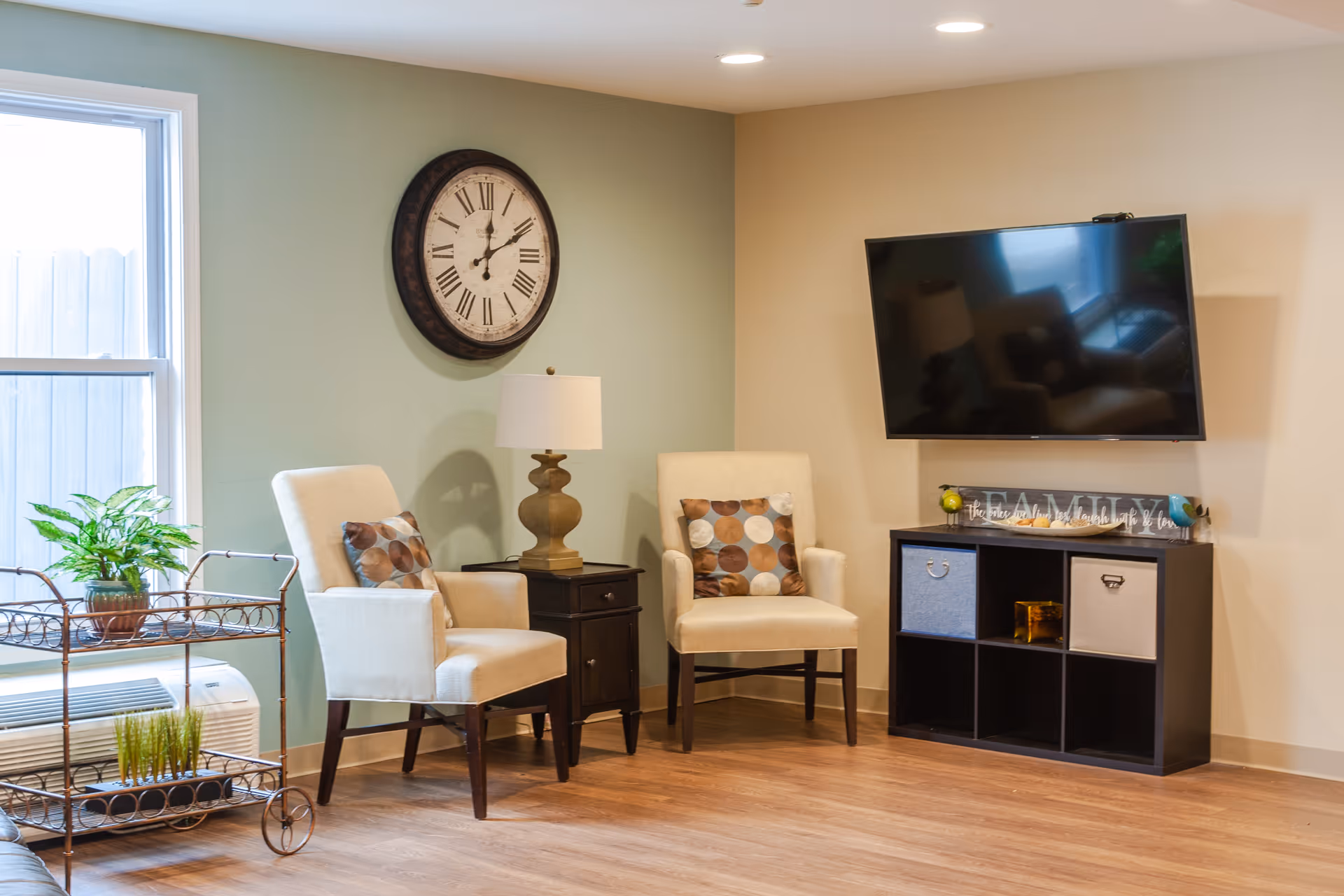 A cozy seating area with two armchairs, a wall clock, a wall-mounted TV, and a small cabinet in a well-lit common room.