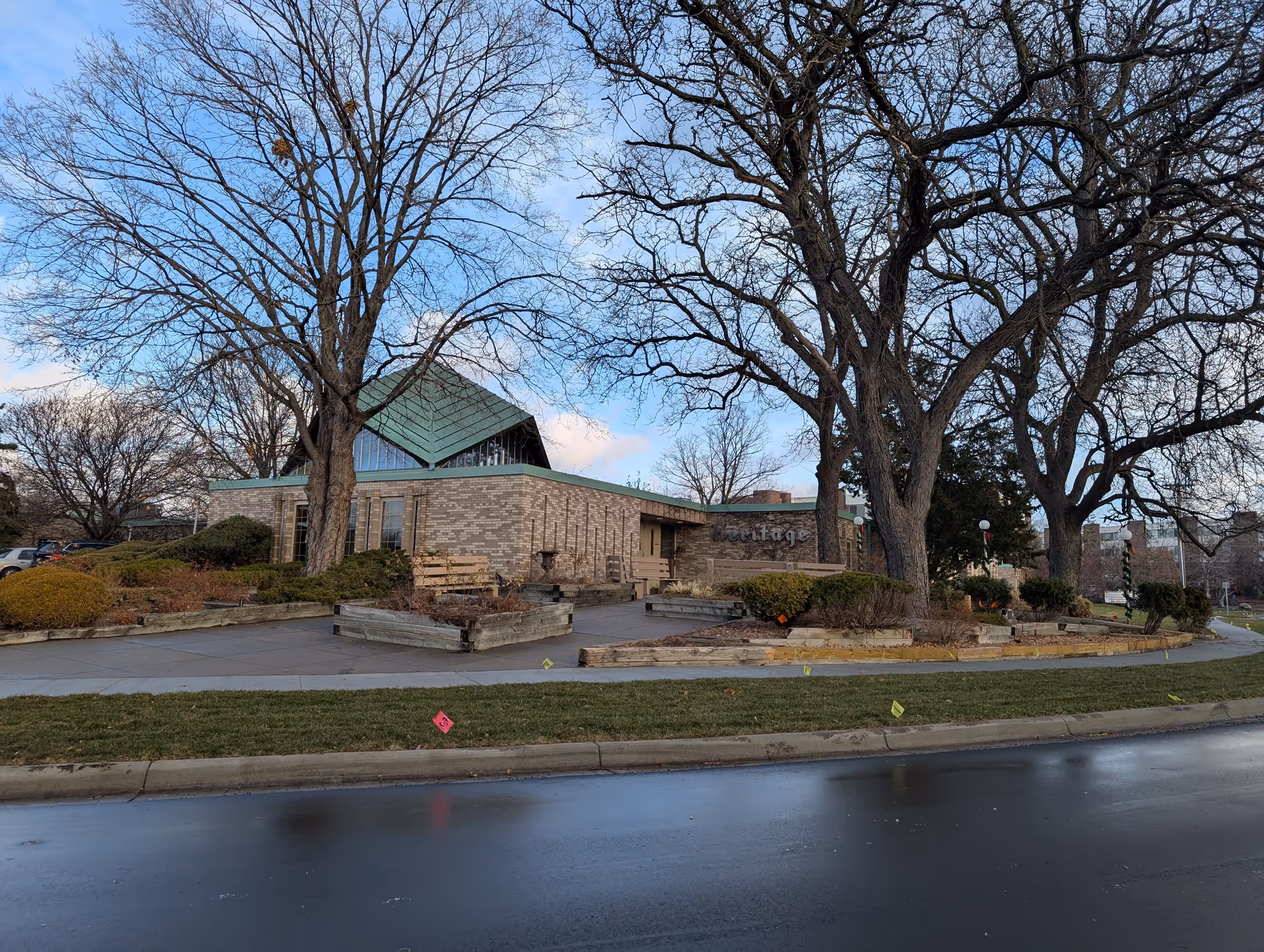 Exterior view of a brick building with a green roof and large leafless trees in front. The building has a sign that reads 'Heritage'. There are benches and landscaped areas with bushes and mulch around the entrance. The sky is partly cloudy and the ground appears wet.