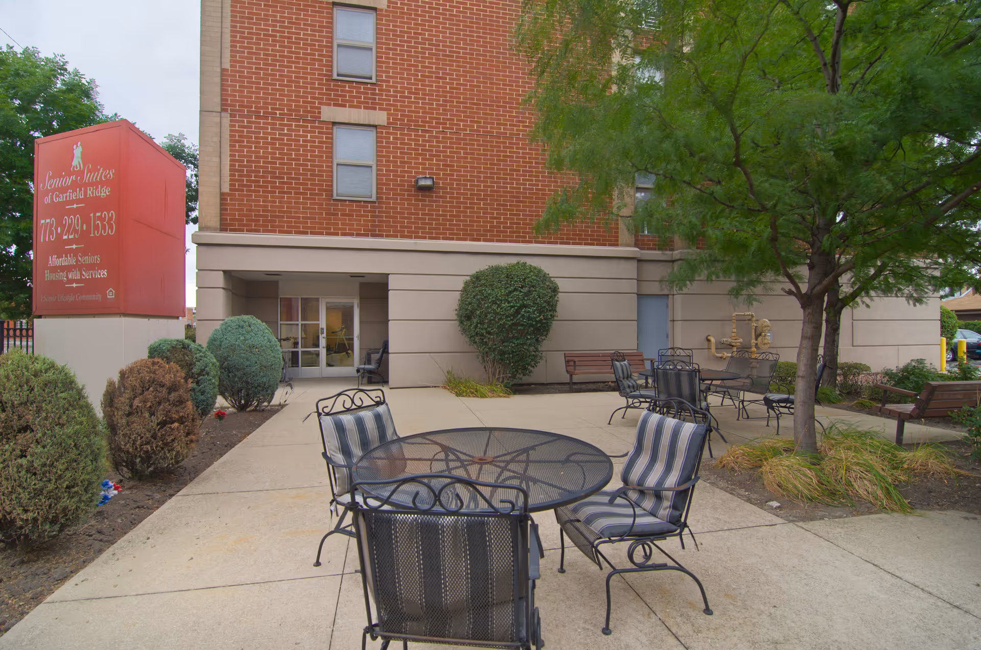 Outdoor patio area at Senior Suites of Garfield Ridge featuring metal tables and chairs with cushions, surrounded by bushes, trees, and a brick building with windows in the background. A large red sign displays the facility name and contact information.