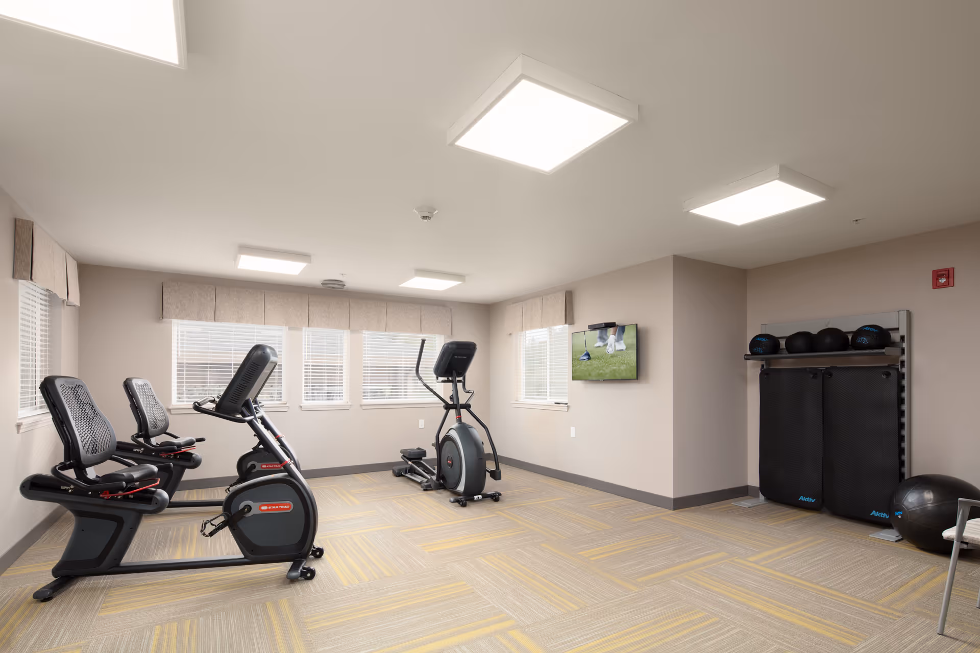 A bright and clean fitness room with exercise equipment including two recumbent bikes and an elliptical machine. The room has beige walls, large windows with blinds, and ceiling lights. On one wall, there is a mounted TV displaying a golf scene. Exercise mats and medicine balls are stored on a rack in the corner.