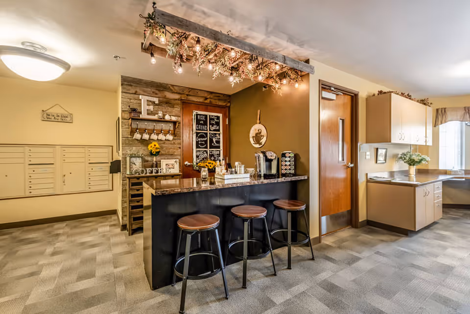 A cozy coffee bar area in a senior living facility with three wooden stools at a counter. The counter has a coffee maker, coffee pods, and decorative items including sunflowers. Above the counter, there is a wooden beam with hanging lights and greenery. To the left, there are mailboxes on the wall and a small sign that reads 'How to be Glad You Are Here'. To the right, there is a door and a built-in desk area with cabinets and a window with curtains.