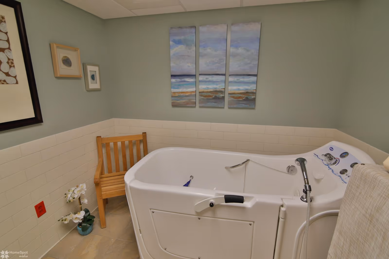 Walk-in accessible bathtub in a tiled bathroom with a wooden chair, wall art, and a small potted plant.