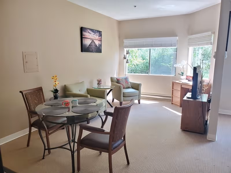 A bright living room with beige walls and carpet, featuring a glass-top round dining table with four chairs, two armchairs with a small side table between them, a wooden TV stand with a flat-screen TV, and a large window with white blinds letting in natural light. A framed picture hangs on the wall above the armchairs.