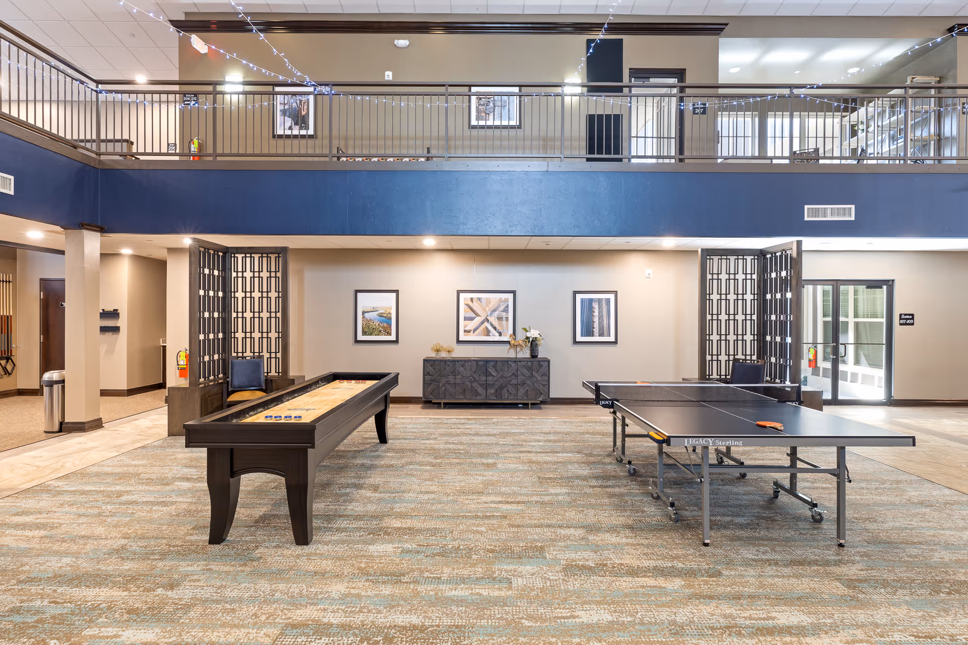 A spacious recreational room in a senior living facility featuring a shuffleboard table and a ping pong table on a patterned carpet. The room has a two-story design with a balcony railing decorated with string lights. The walls are adorned with framed artwork, and there is a dark wooden cabinet against the far wall. The space is well-lit with recessed lighting and natural light coming through glass doors on the right.
