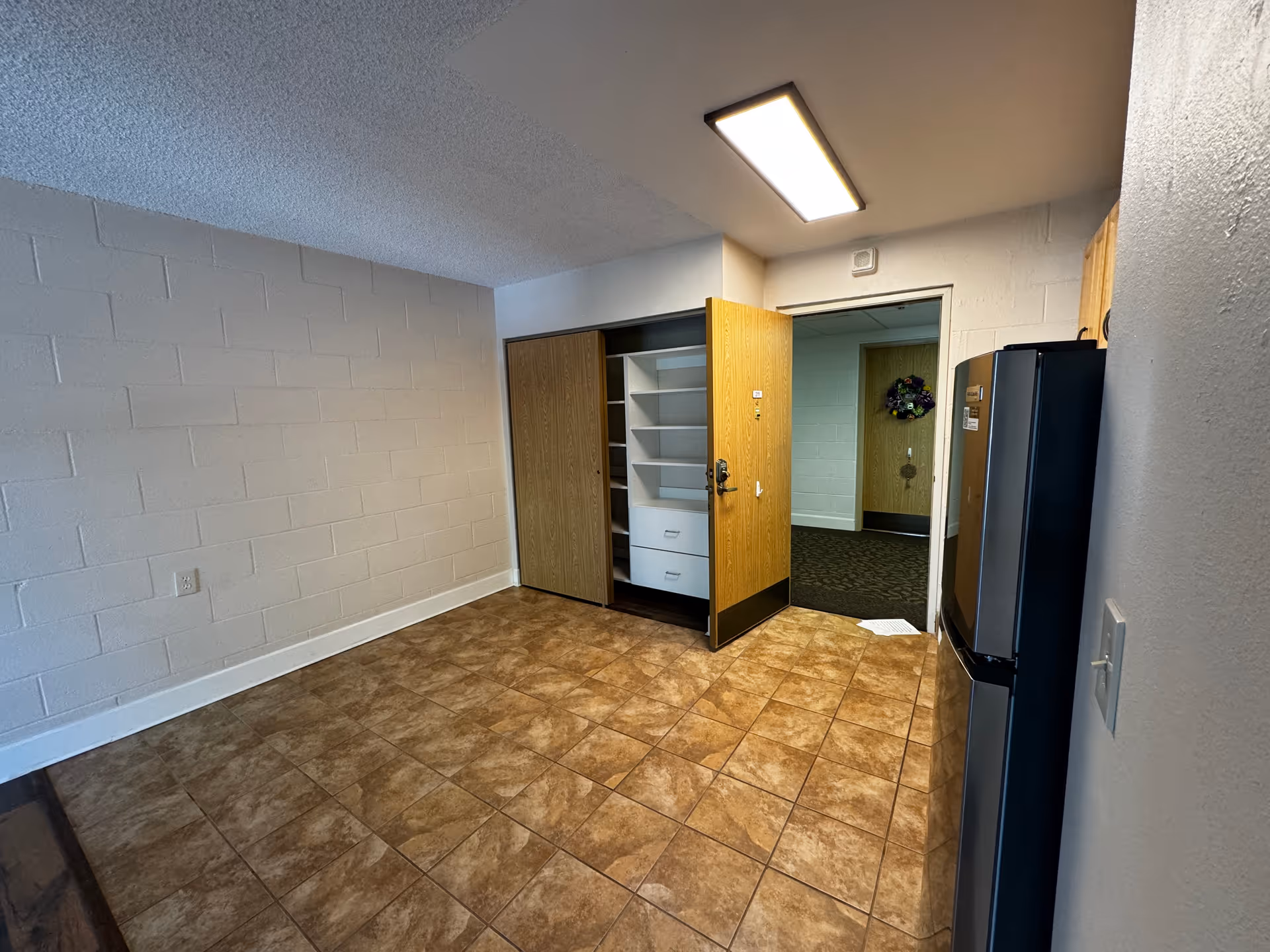 An empty room with tiled floor and white painted cinder block walls. There is a closet with wooden sliding doors, one door open revealing shelves and drawers inside. A wooden door is open showing a hallway with carpet and a wreath on another door. A refrigerator is visible on the right side of the image. The ceiling has a rectangular fluorescent light fixture.