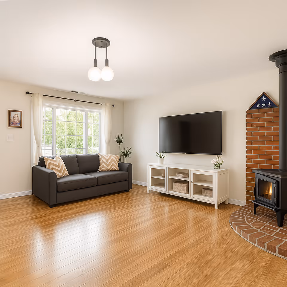 A bright living room with a large window covered by white curtains, a dark gray sofa with two beige and white chevron-patterned pillows, a white TV stand with a flat-screen TV mounted on the wall above it, and a black wood-burning stove set on a brick hearth with a folded American flag displayed above.