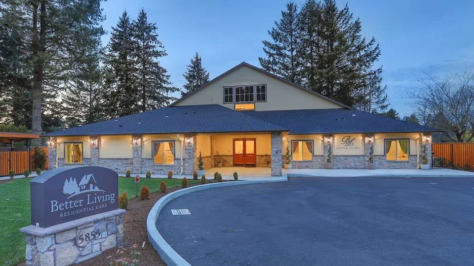 Exterior front view of Better Living Residential Care facility at dusk, showing a single-story building with stone accents, lit windows, and a curved driveway. A sign in front reads 'Better Living Residential Care' with an address number 5851. Tall trees surround the property.