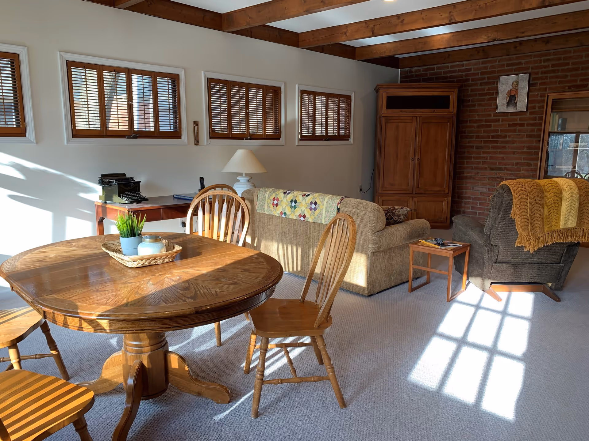 Sunlit living room and dining area with a round wooden table and chairs, sofas, a wooden armoire, and exposed ceiling beams.