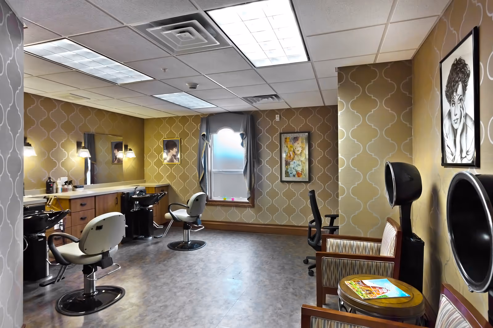 Interior view of a hair salon area in a senior living facility with two salon chairs in front of sinks and mirrors, patterned wallpaper, a window with curtains, and two hair drying stations next to a small seating area with chairs and a table holding magazines.