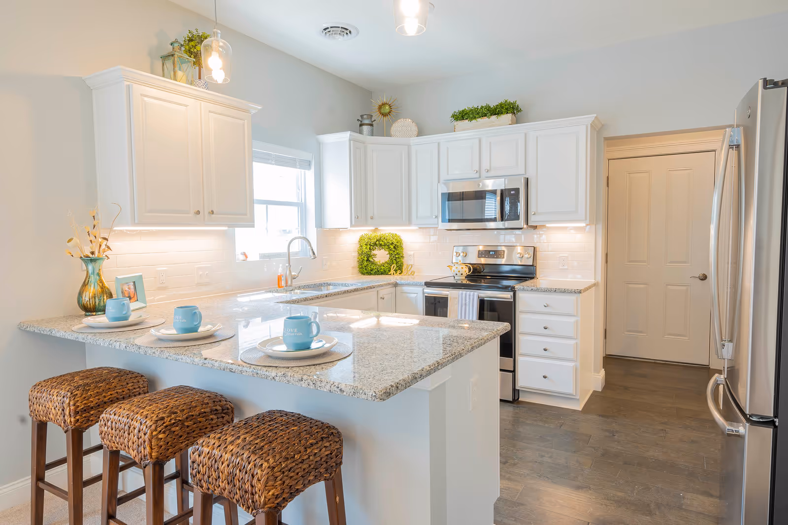 Bright and modern kitchen with white cabinets, granite countertops, and stainless steel appliances including a refrigerator, stove, and microwave. Three woven bar stools are placed at the kitchen island, which is set with plates and blue mugs. The kitchen has under-cabinet lighting, a window above the sink, and decorative plants and items on top of the cabinets.