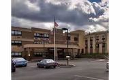 Exterior view of a multi-story building with a beige facade under a cloudy sky. There is a flagpole with an American flag in front of the building and several parked cars in the parking lot.