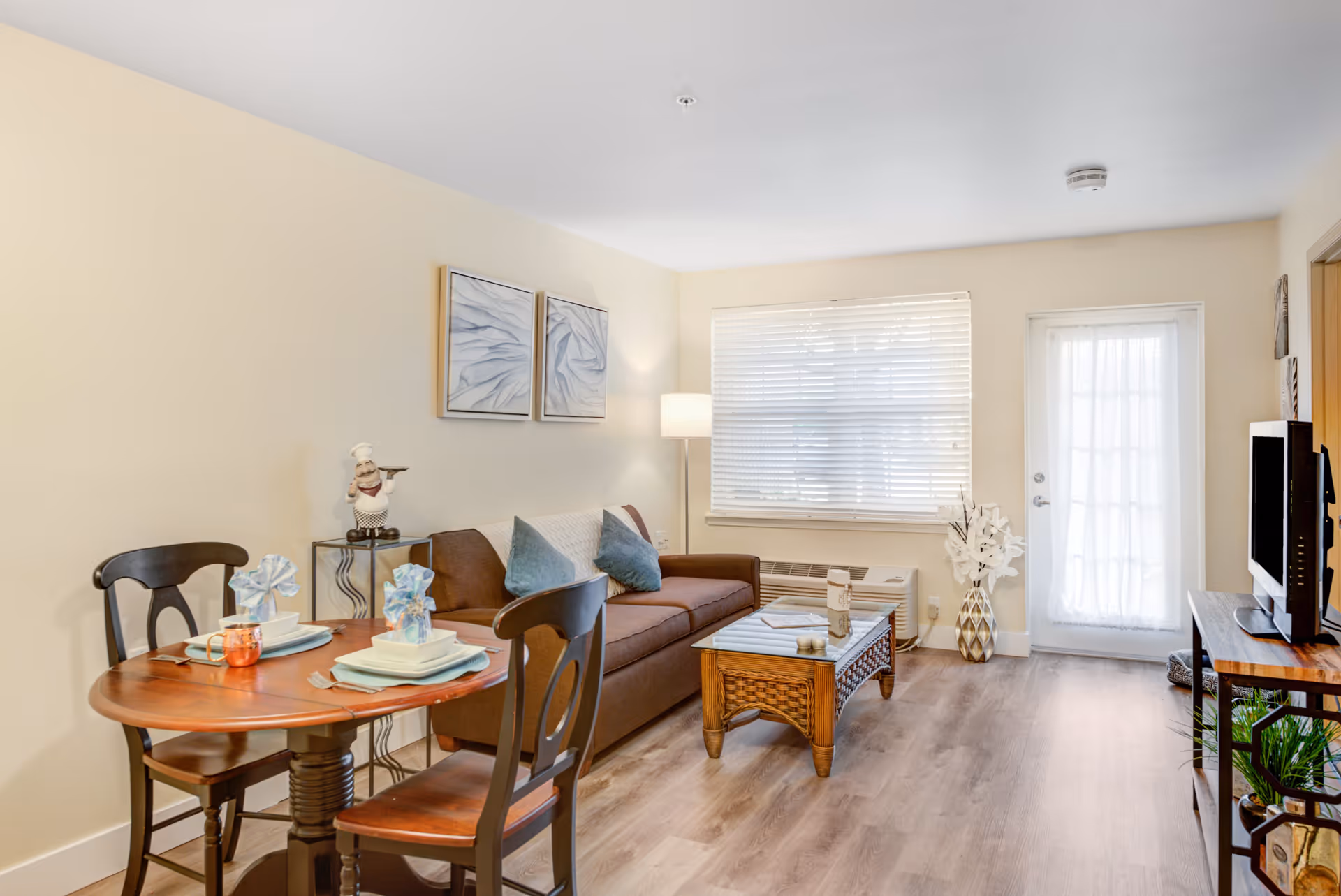 A bright living room with a brown sofa adorned with two blue cushions, a wooden coffee table, and a small round dining table set for two with plates, napkins, and cups. The room has light-colored walls, a large window with blinds, a floor lamp, a TV on a stand, and a door with a sheer curtain. Decorative items include wall art, a small chef figurine on a side table, and a vase with white flowers.