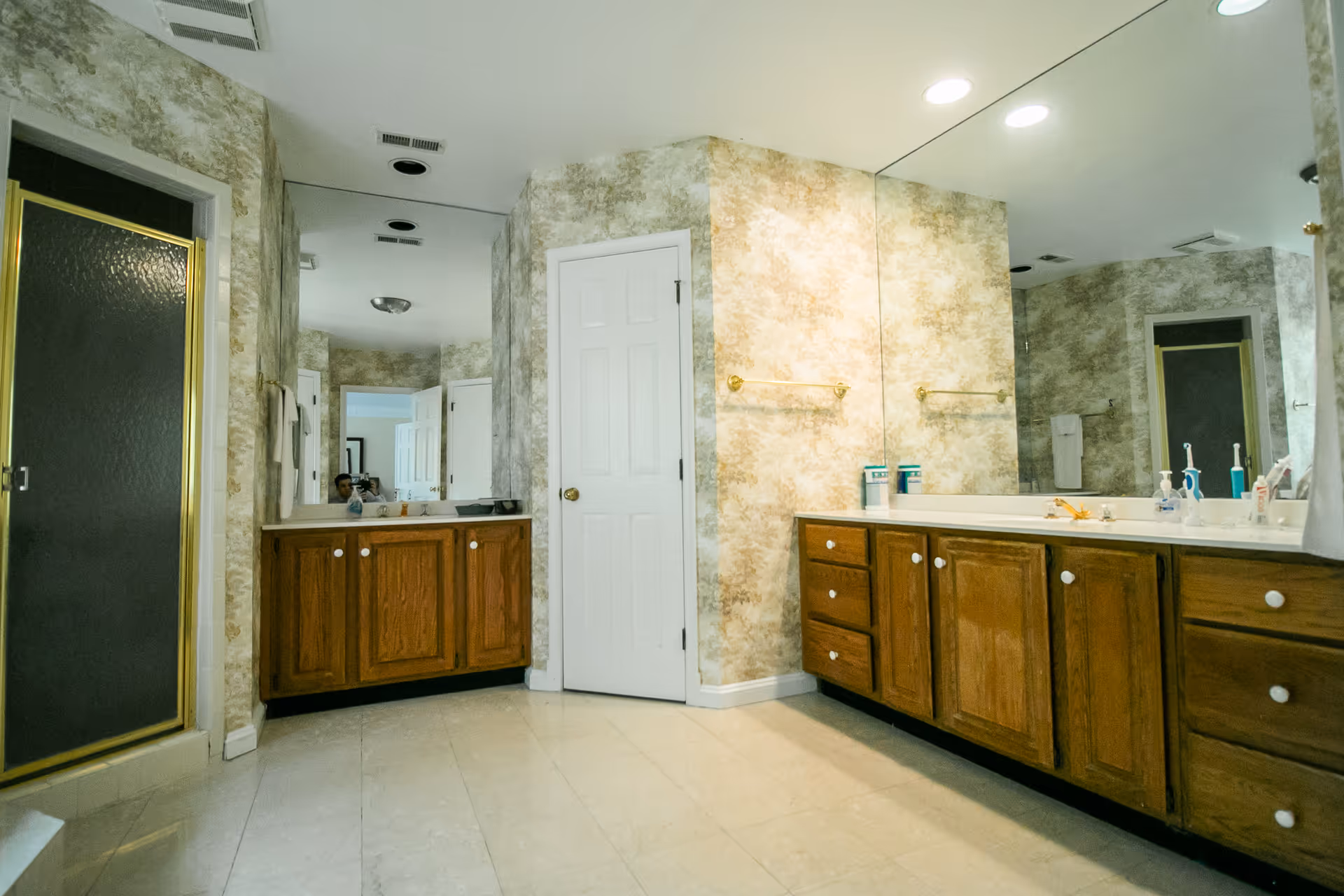 A spacious bathroom with beige patterned wallpaper, two wooden vanities with white countertops and sinks, a large mirror covering the wall above the vanities, a white door in the center, and a shower with a frosted glass door and gold trim on the left side.