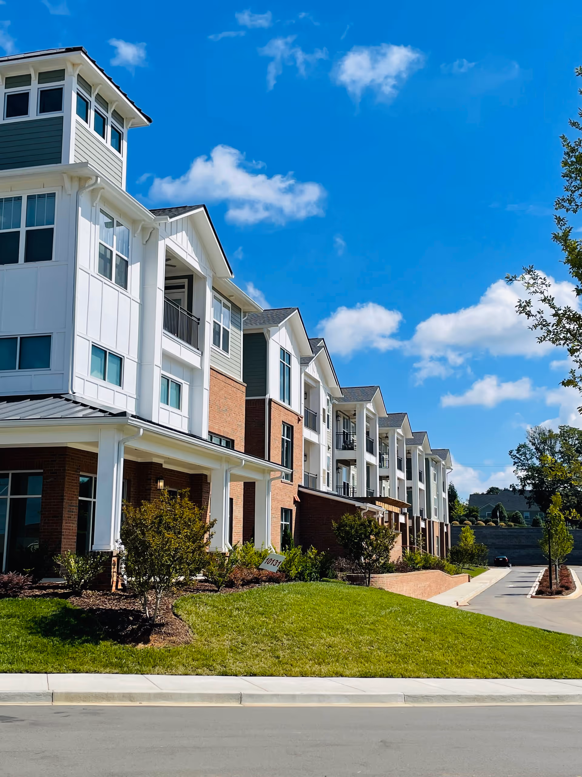 Front exterior of a modern multi-story senior living building with balconies, landscaped lawn, and a bright blue sky.
