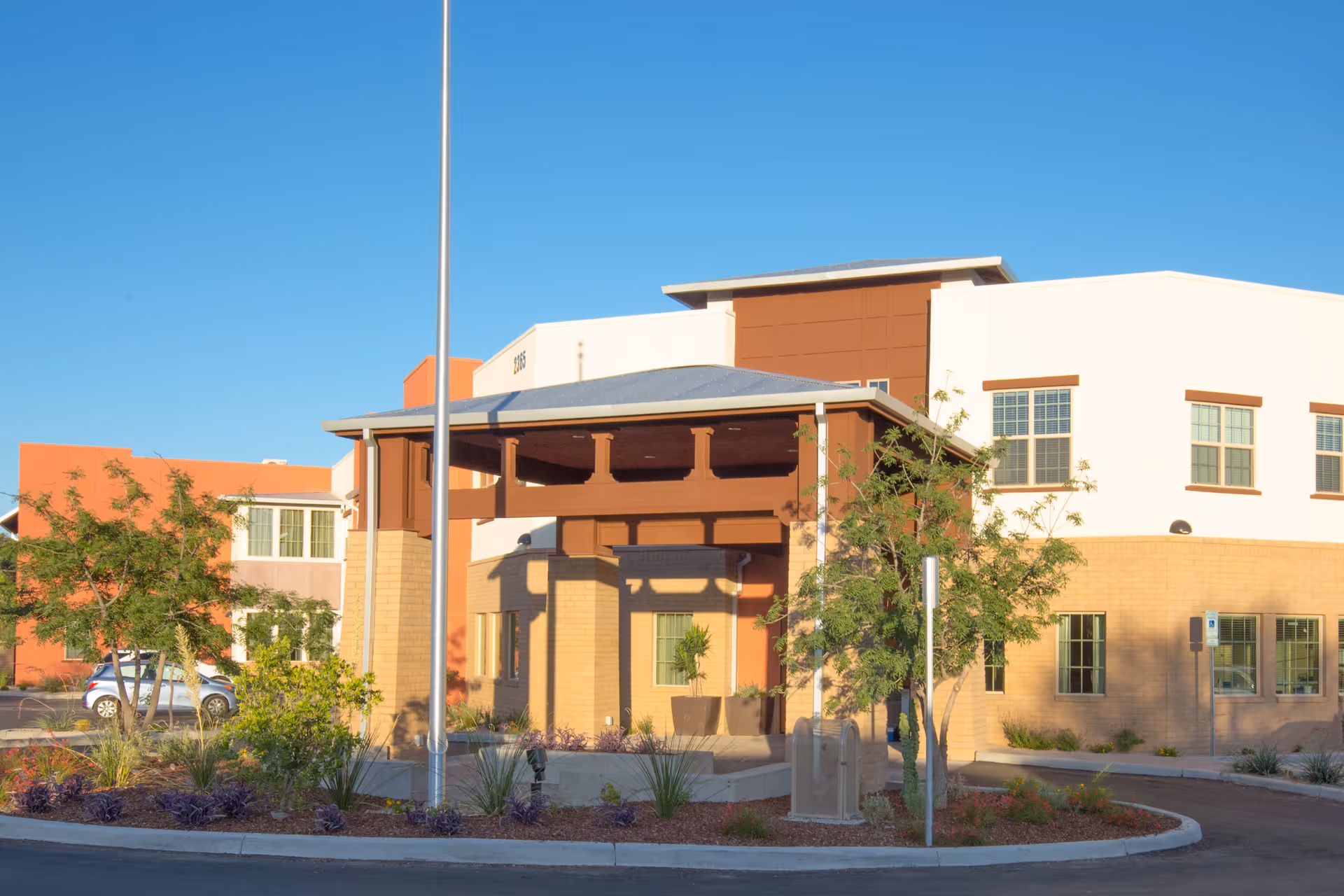 Exterior view of a senior living facility building with a covered entrance, beige and brown walls, several windows, and landscaping including trees and shrubs under a clear blue sky.