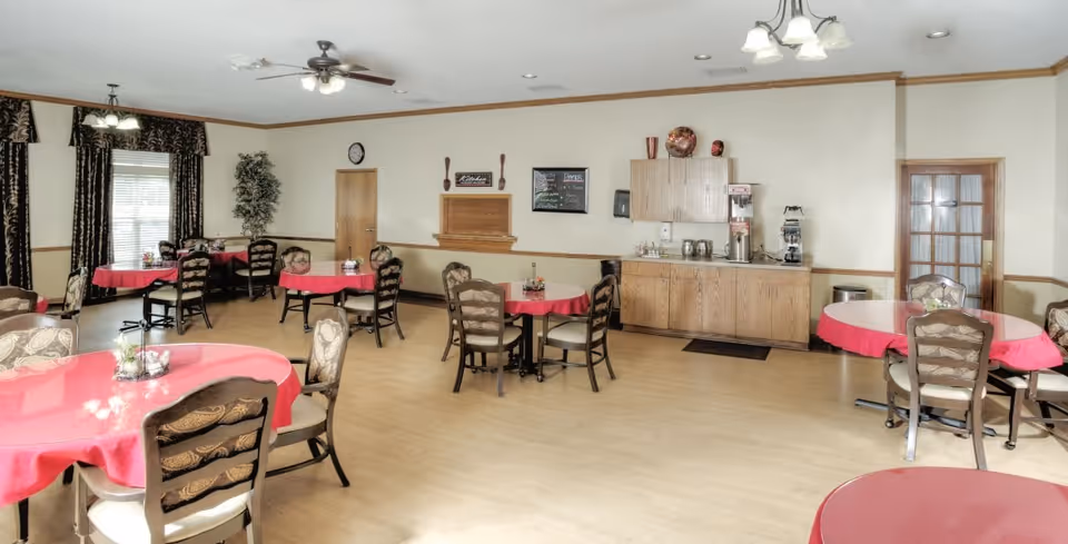 A dining room with multiple round tables covered with red tablecloths and surrounded by chairs. The room has light-colored walls with wood trim, a wooden cabinet with a coffee and beverage station, a ceiling fan, and windows with dark patterned curtains. The floor is light wood, and there are ceiling lights providing illumination.
