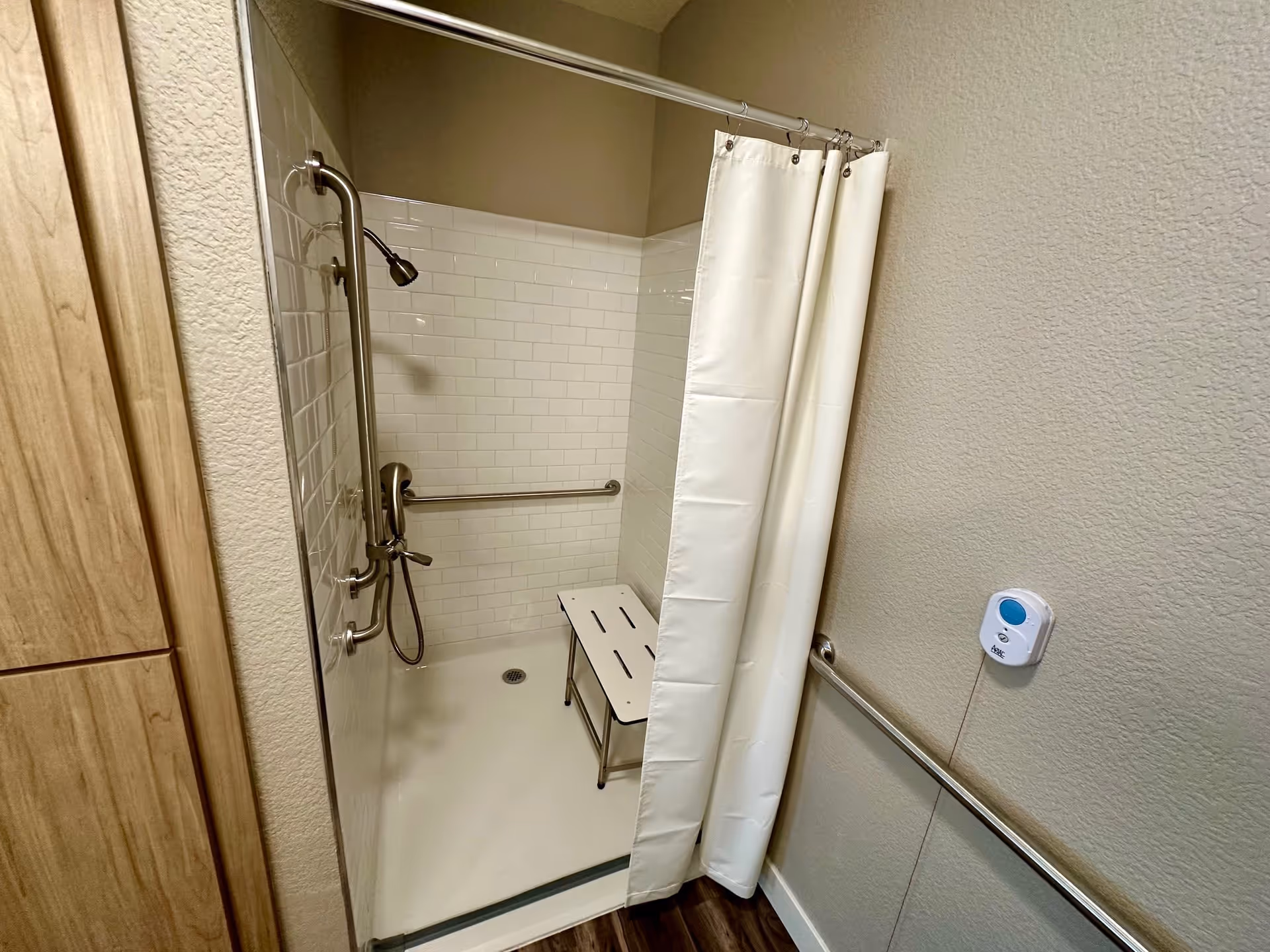 A shower area with white tiled walls, a white shower curtain, a metal grab bar, a handheld showerhead, and a white shower bench. The floor outside the shower is wood-patterned, and there is a call button on the wall.