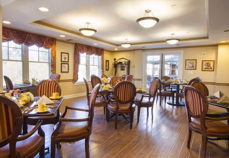 A bright dining room with multiple round tables set with yellow cups, saucers, and folded napkins. The room has wooden floors, large windows with red and gold curtains, and warm yellow walls adorned with framed pictures. There are elegant wooden chairs with red and gold striped upholstery around the tables. Ceiling lights provide soft illumination, and glass doors lead to an outdoor area.