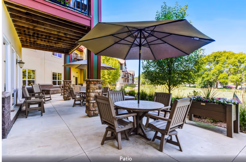 Outdoor patio area at The Homestead at Anoka featuring multiple round tables with umbrellas and wooden chairs. The patio is adjacent to a building with stone pillars and overlooks a green space with trees and plants.
