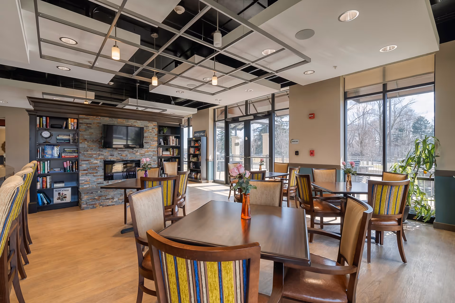 A bright and spacious common area with multiple wooden tables and chairs, each table decorated with a small vase of pink flowers. The room features large windows letting in natural light, a stone fireplace with a mounted flat-screen TV above it, and bookshelves filled with books on either side. The ceiling has a modern design with hanging lights, and there are plants near the windows.
