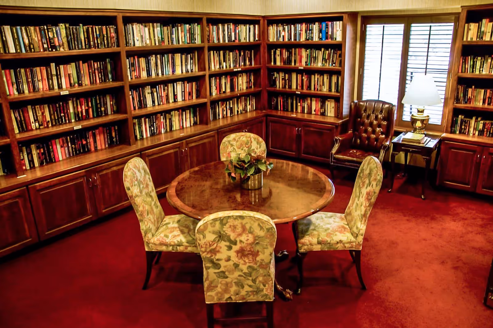 Cozy library-style room with wall-to-wall bookshelves, a round wooden table surrounded by four floral-upholstered chairs, and a leather armchair by a window.