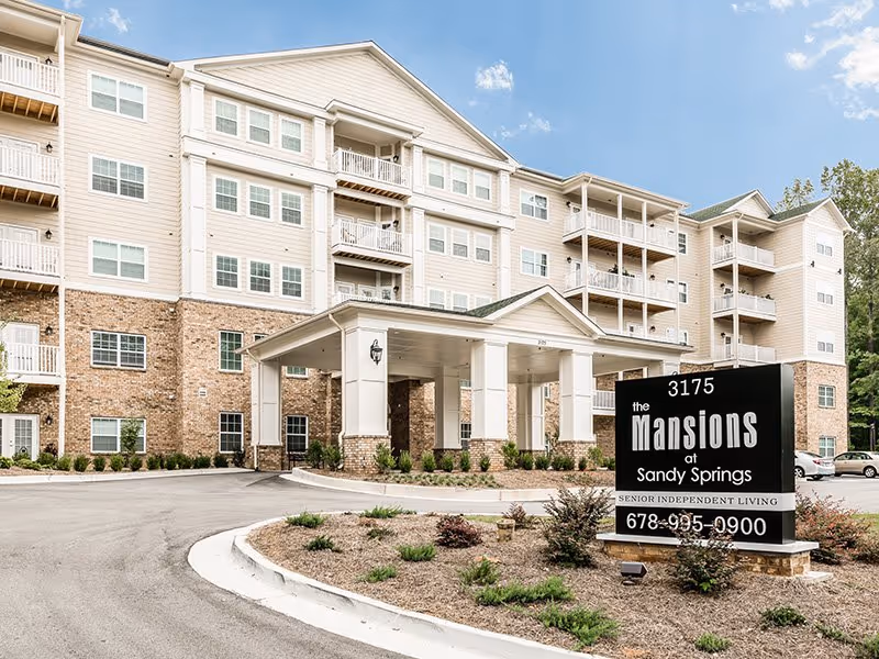 Exterior front view of The Mansions at Sandy Springs Senior Independent Living building with a covered entrance, multiple balconies, and a sign displaying the facility name and contact number.