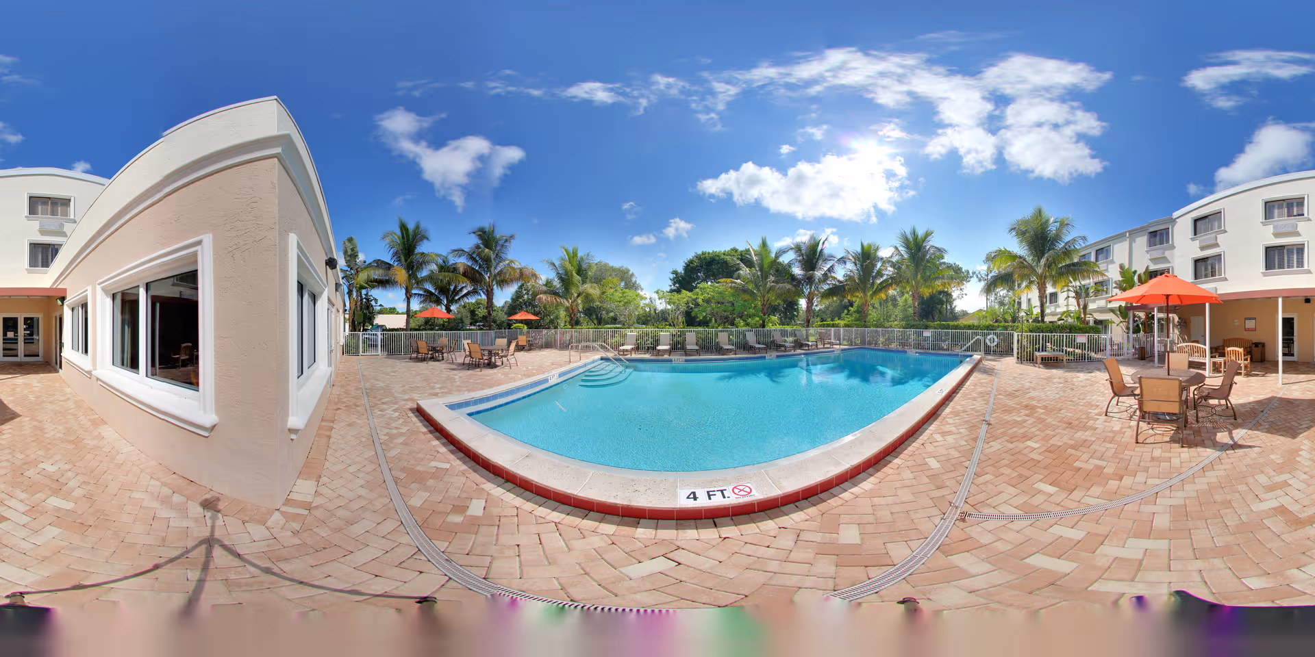 Outdoor swimming pool area at Tuscany Villa of Naples with clear blue water, surrounded by a brick-paved deck. Several tables with chairs and red umbrellas are placed around the pool. Palm trees and greenery are visible in the background under a bright blue sky with some clouds. The building of the facility is visible on both sides of the pool area.
