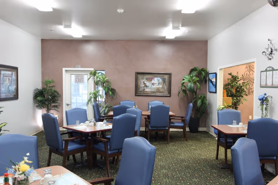 A dining room in an assisted living facility with several wooden tables and blue cushioned chairs arranged neatly. The room has a green patterned carpet, beige and white walls, framed artwork, and several potted plants. There is a menu board on one wall and bright overhead lighting.