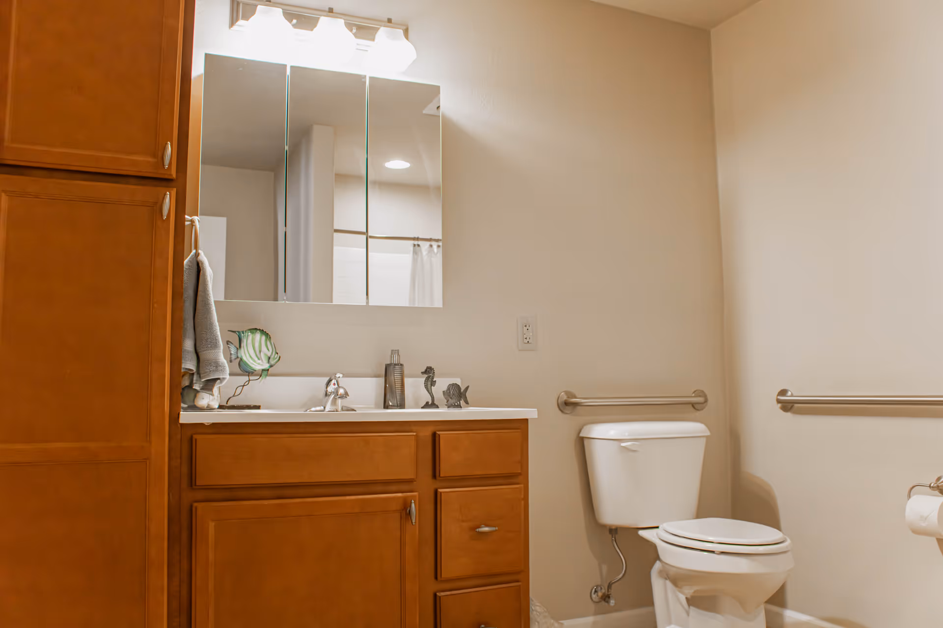 A bathroom with wooden cabinets, a white sink with a silver faucet, a mirror with three panels above the sink, and a toilet with grab bars on the walls for accessibility. There is a towel hanging on the side of the cabinet and decorative items on the sink counter.