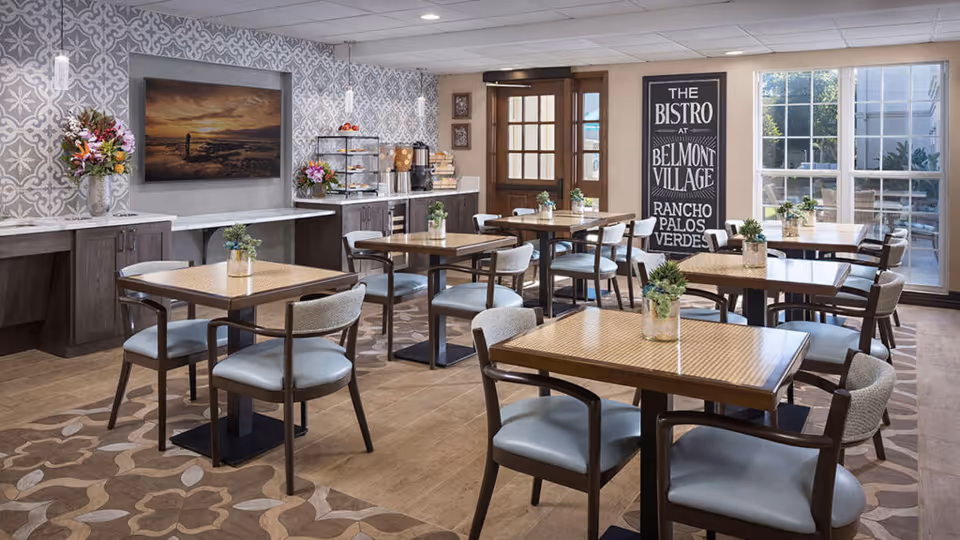 A bright and inviting dining area at Belmont Village Senior Living Rancho Palos Verdes featuring several wooden tables with chairs upholstered in light blue. Each table has a small plant centerpiece. The room has patterned wallpaper, a large window letting in natural light, a door leading outside, and a sign that reads 'The Bistro at Belmont Village Rancho Palos Verdes'. There is a counter with coffee and pastries on the left side.