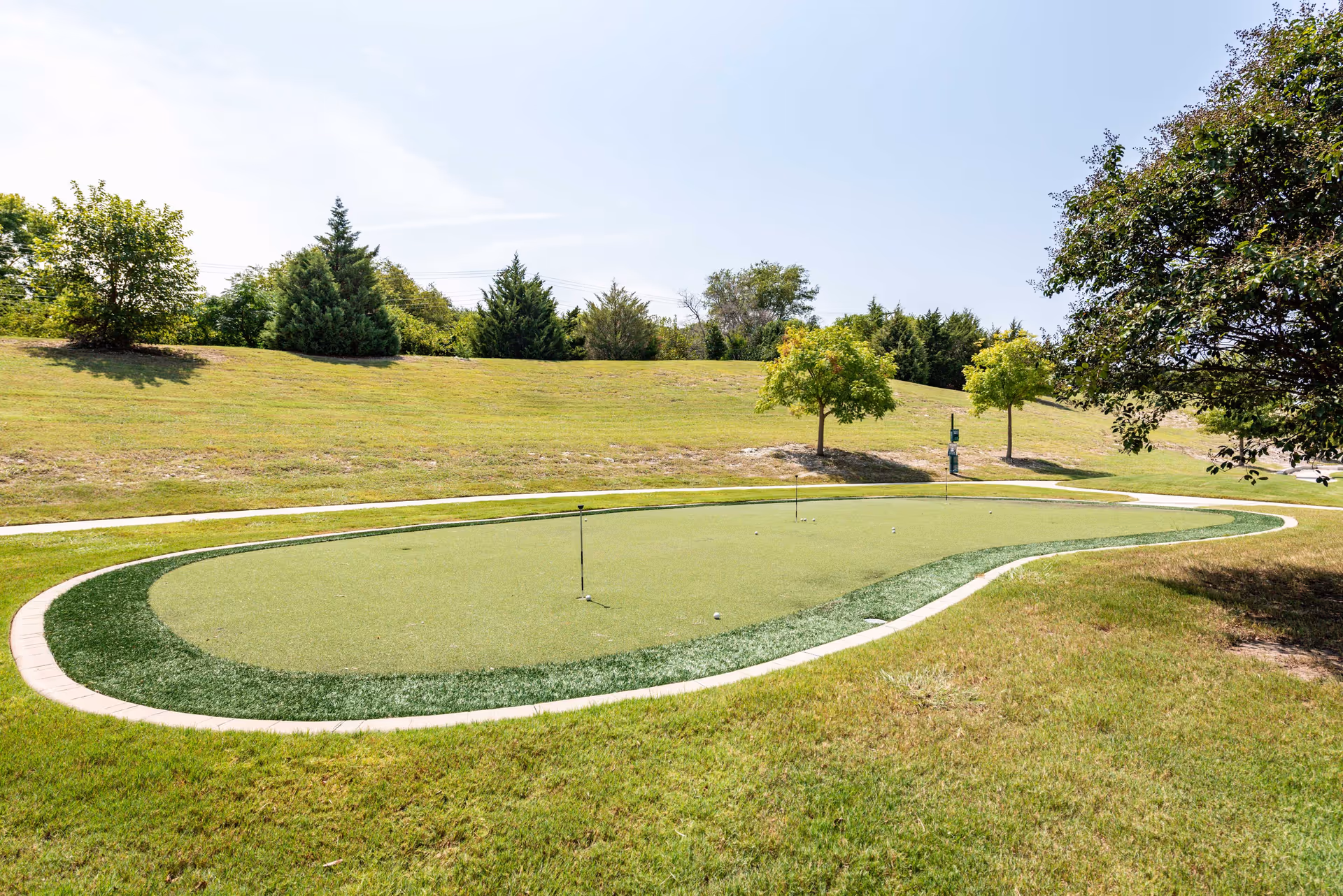 A small outdoor putting green surrounded by grass, trees, and a walking path under a clear sky.