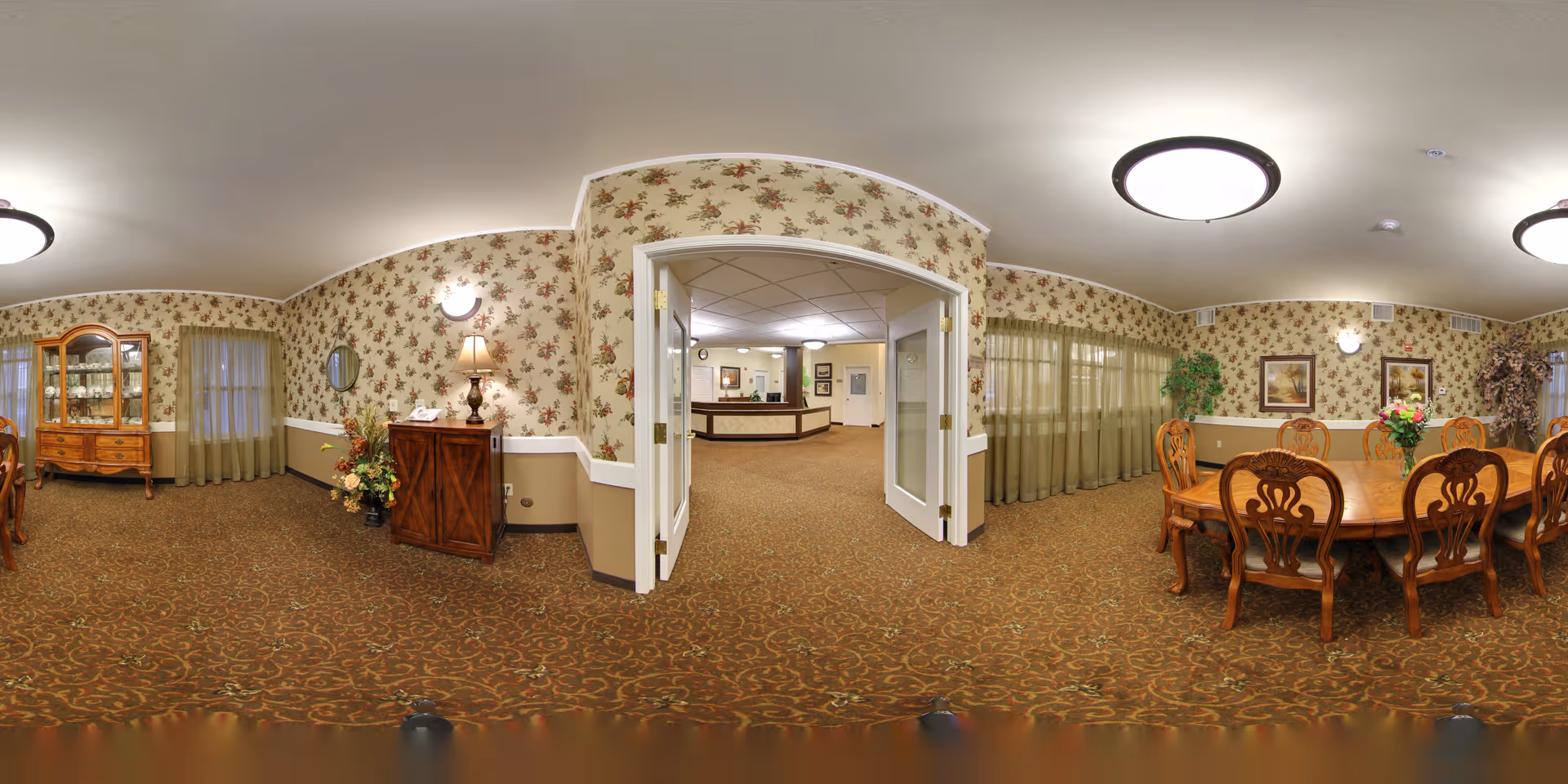 Panoramic interior of a senior living dining room with a large wooden table and chairs, floral wallpaper, a china cabinet and an open doorway to a reception area.