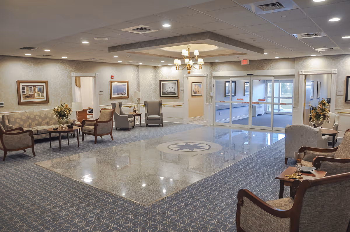 A spacious and well-lit lobby area with polished marble flooring in the center and patterned carpet around the edges. The room features several upholstered chairs and sofas arranged in seating groups, side tables with lamps and flower arrangements, framed artwork on the walls, and a chandelier hanging from a recessed ceiling. Large glass double doors lead to an outside area.