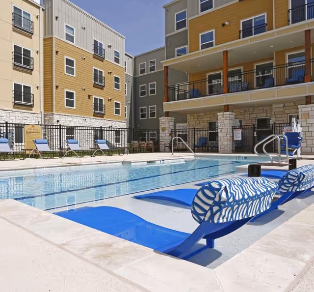 Outdoor swimming pool area at Sansom Pointe Senior facility with blue lounge chairs partially submerged in the shallow end of the pool. Surrounding the pool are multiple buildings with balconies and windows, and additional lounge chairs lined up along the poolside under a clear sky.