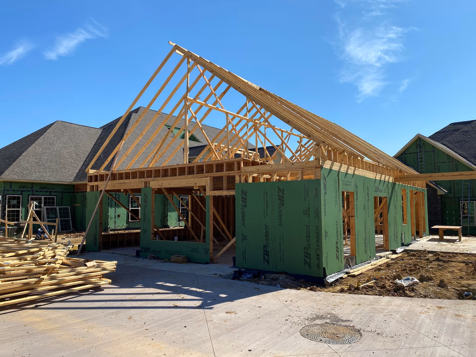 A building under construction with wooden framing and green sheathing panels. The roof structure is partially completed with exposed wooden rafters. The surrounding area has construction materials and a concrete surface under a clear blue sky.
