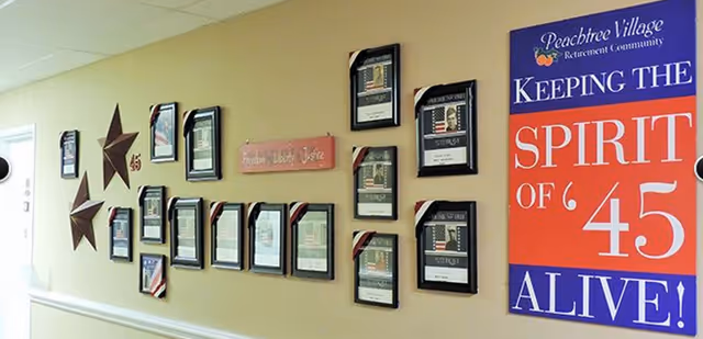 Interior wall of Peachtree Village Retirement Community decorated with framed documents and plaques arranged in a grid pattern, two large decorative stars, and a large colorful sign that reads 'Peachtree Village Retirement Community KEEPING THE SPIRIT OF '45 ALIVE!'