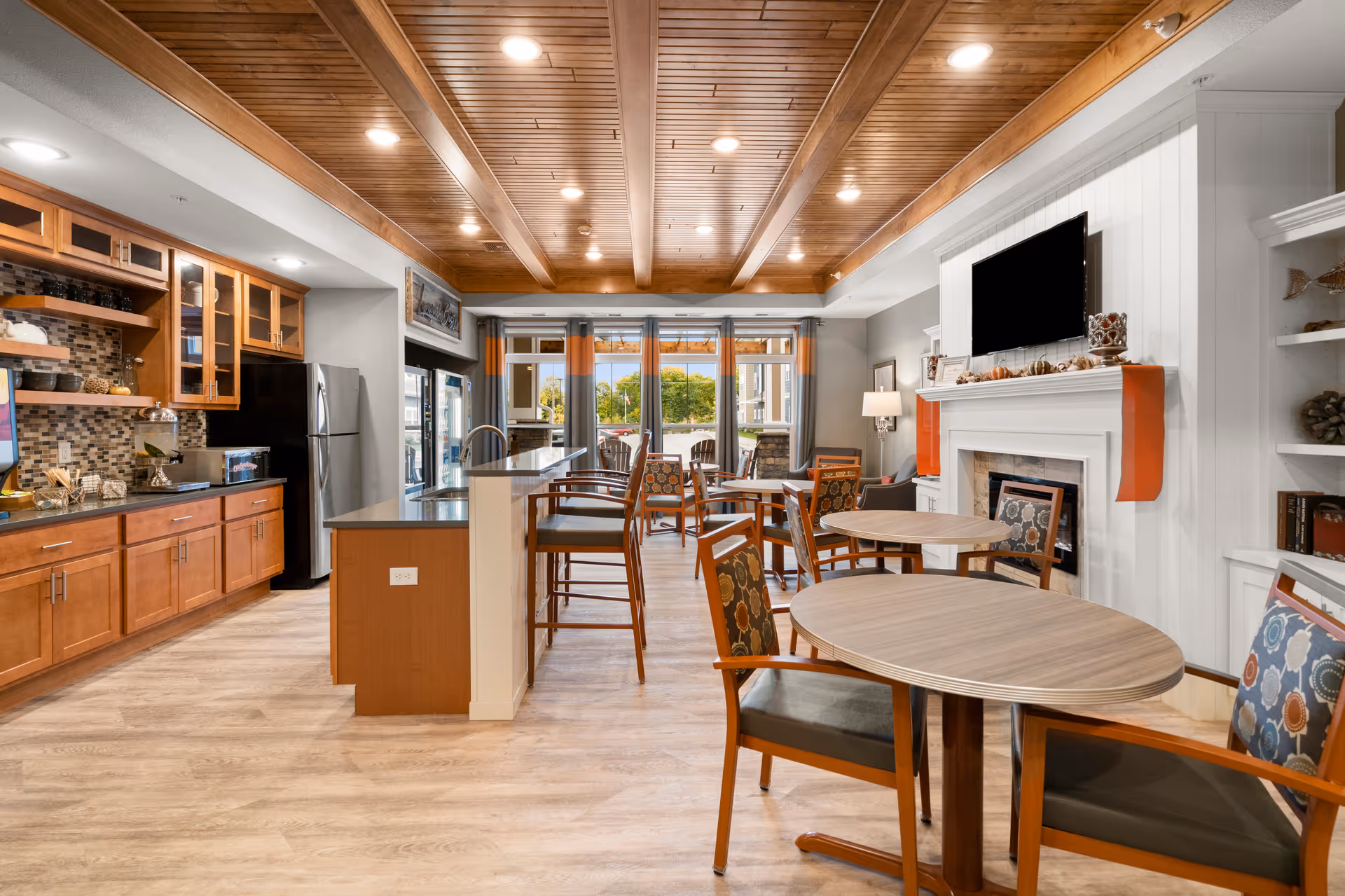 A bright and spacious common area in a senior living facility featuring a modern kitchen with wooden cabinets and stainless steel refrigerator on the left, a central island with bar stools, and multiple round tables with chairs arranged for dining or socializing. The room has a wooden beamed ceiling with recessed lighting, large windows with curtains letting in natural light, and a white fireplace with a mounted flat-screen TV above it on the right side.