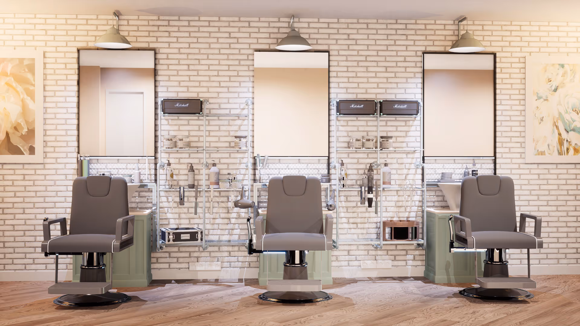 A modern salon area with three gray barber chairs in front of a white brick wall. Each station has a large mirror, shelves with various hair care products, and mounted lights above. The floor is wooden, and there are floral paintings on the walls on either side.
