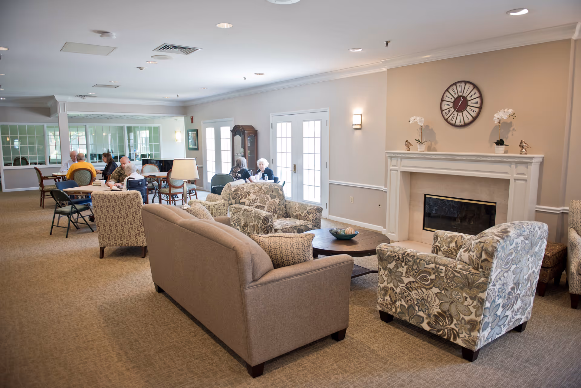 A spacious senior living common area with comfortable seating including a beige sofa and patterned armchairs arranged around a coffee table in front of a fireplace. Several elderly people are seated at tables in the background, engaging in conversation. The room has beige walls, carpeted floor, and large windows with French doors letting in natural light.