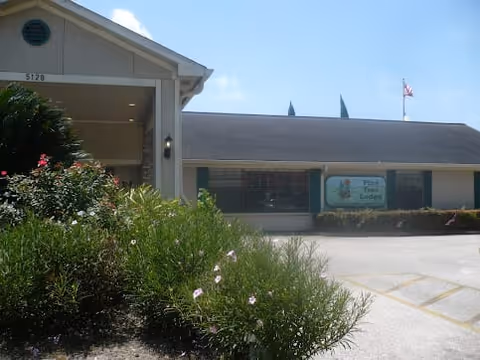 Exterior view of a single-story building with a sign that reads 'Pine Tree Lodge'. The building has a light-colored facade with a covered entrance and some bushes and flowers in the foreground. The sky is clear with a few clouds.