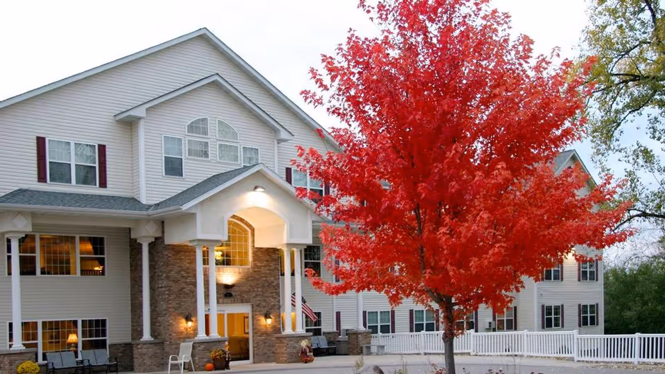 Exterior view of a senior living facility building with white siding and brick accents, featuring multiple windows and a covered entrance with columns. In front of the building, there is a vibrant red tree and a white fence surrounding the area.