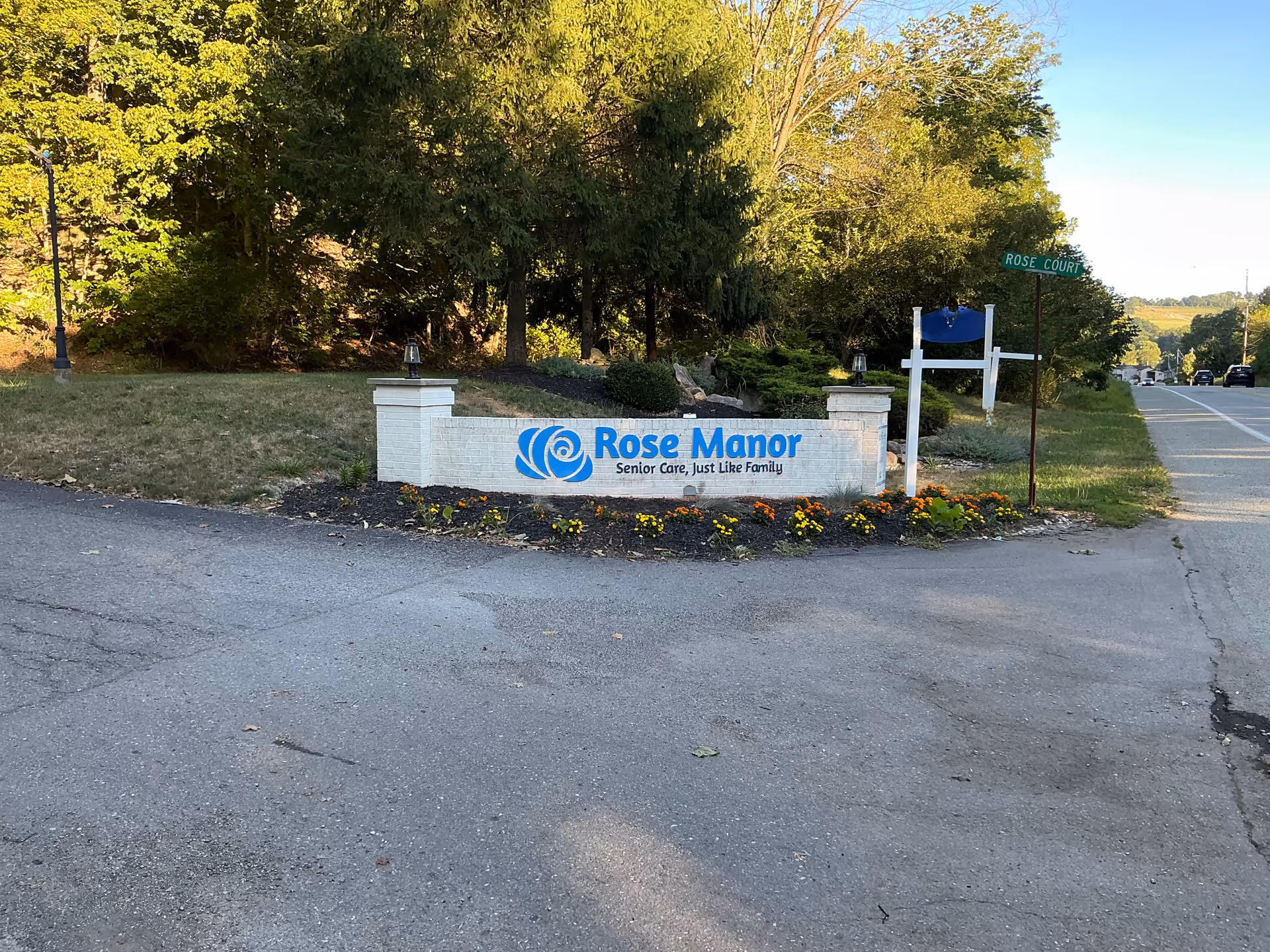 A white brick entrance sign for Rose Manor surrounded by flowers and trees beside a roadway.