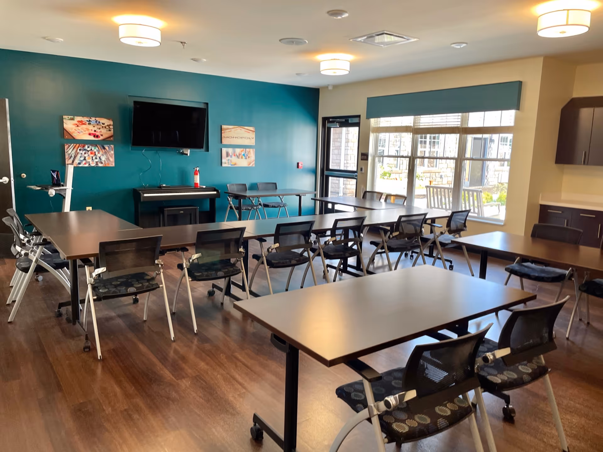 A well-lit room with multiple tables arranged in a U-shape and several chairs around them. The room has wooden flooring, a teal accent wall with a mounted flat-screen TV, and three framed pictures. There is a piano beneath the TV and a fire extinguisher on top of it. Large windows on one side let in natural light, and there are ceiling lights providing additional illumination. Cabinets and a countertop are visible on the right side of the room.