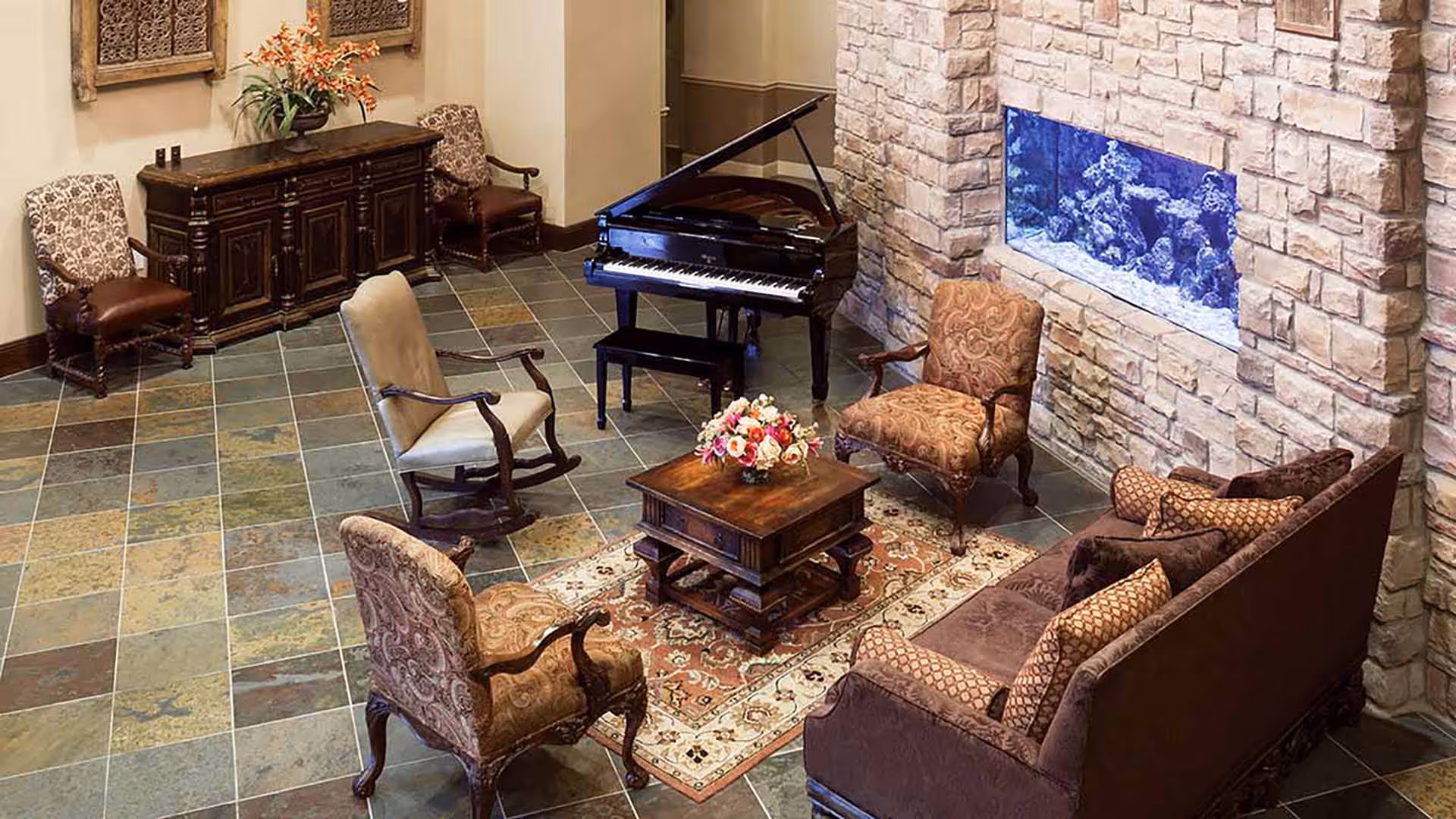 A cozy living room area with a stone wall featuring a built-in aquarium, a grand piano, a wooden coffee table with a flower arrangement, a patterned rug, a sofa with multiple cushions, and several upholstered armchairs arranged around the coffee table.