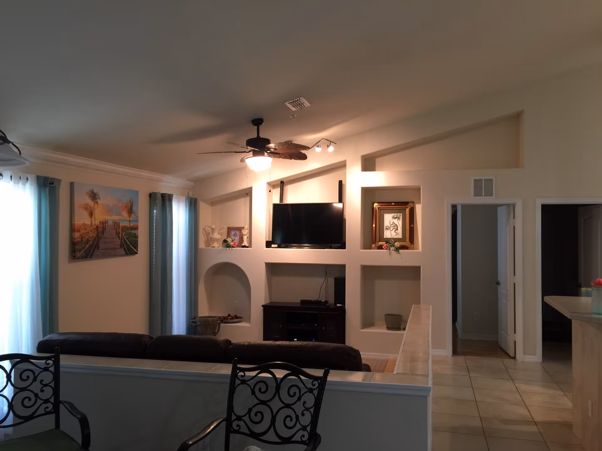 Interior view of a living room with a ceiling fan and light fixture, a wall-mounted flat screen TV, decorative shelves with framed pictures and vases, a brown leather sofa, two wrought iron chairs with green cushions, tiled floor, and windows with teal curtains allowing natural light.