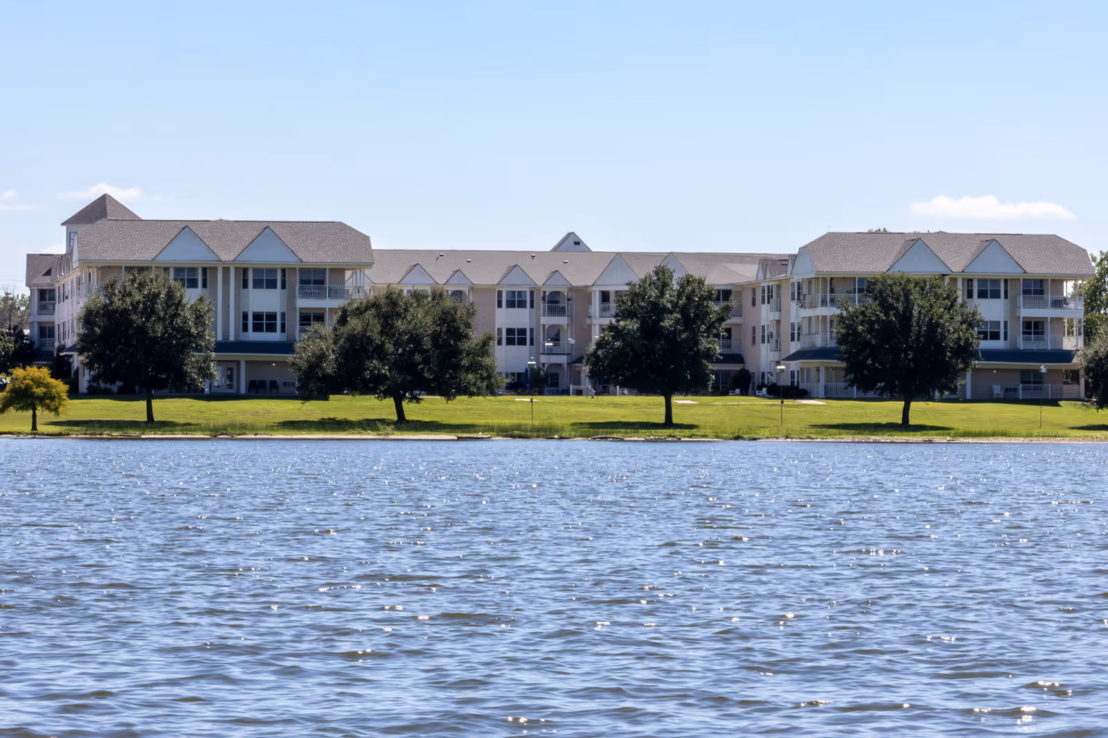 View of a large senior living facility building named Lakestone Terrace Senior Living, seen across a body of water with a grassy area and several trees in front of the building under a clear blue sky.