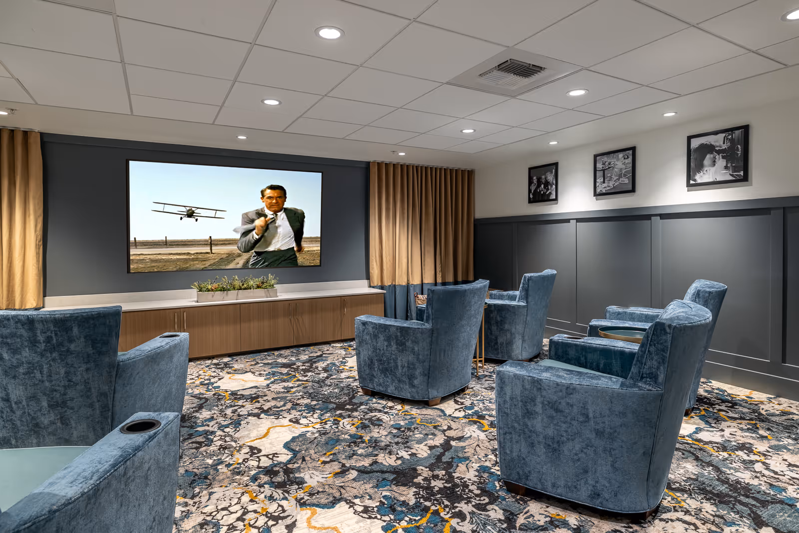 A cozy movie room with six blue velvet armchairs arranged facing a large flat-screen TV mounted on a dark gray wall. The TV is showing a scene from a classic movie with a man running and a biplane flying behind him. The room has a patterned carpet with blue, gray, and yellow tones, beige curtains on either side of the TV, and three black-and-white framed photos on the wall to the right.