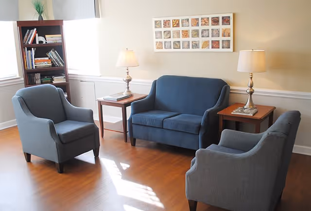 A bright seating area with a blue loveseat and matching armchairs, side tables with lamps, a bookshelf, and framed wall art.