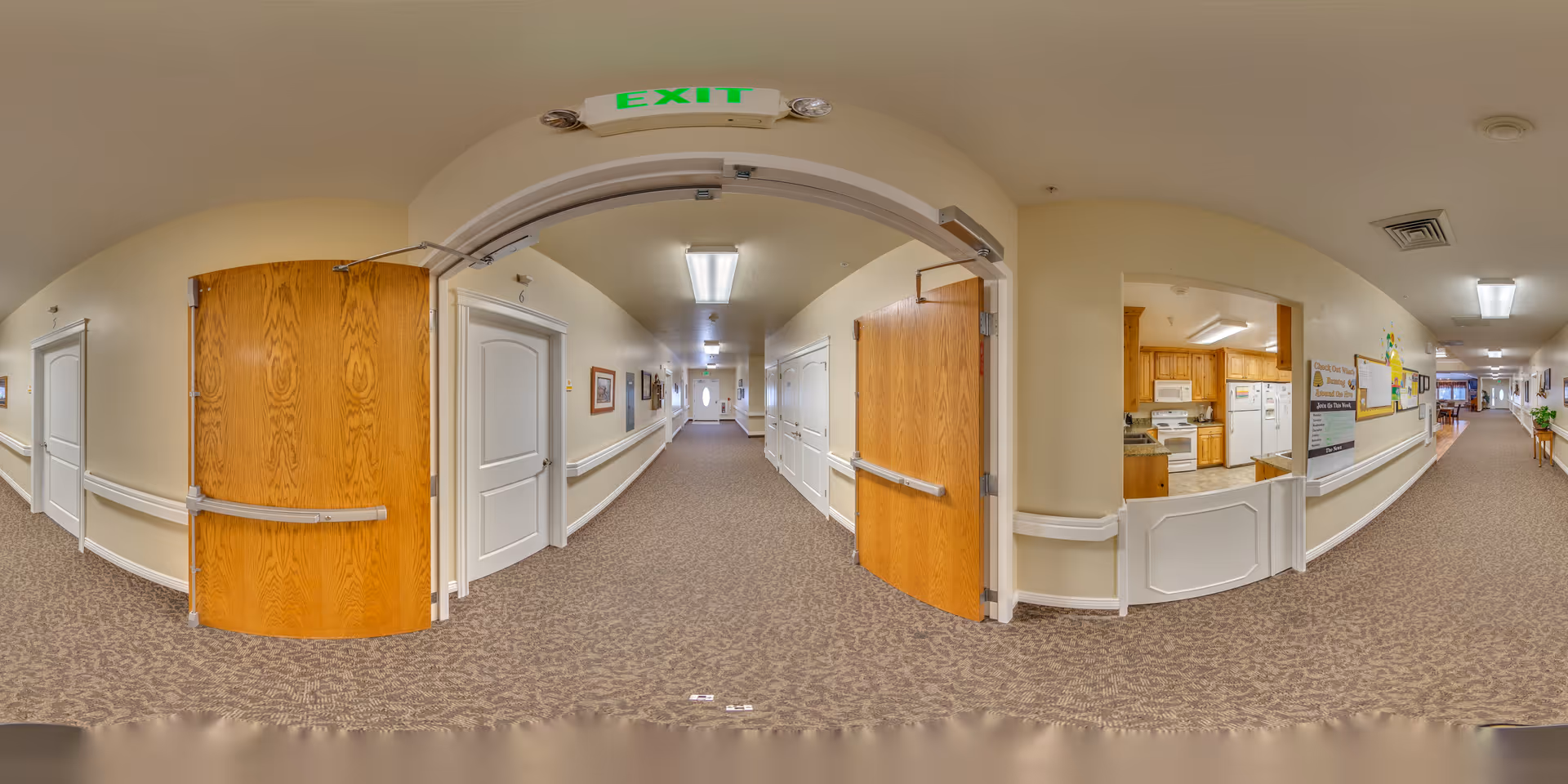 Carpeted interior hallway of a senior living facility with open wooden double doors, an EXIT sign overhead, and a kitchen service window on the right.
