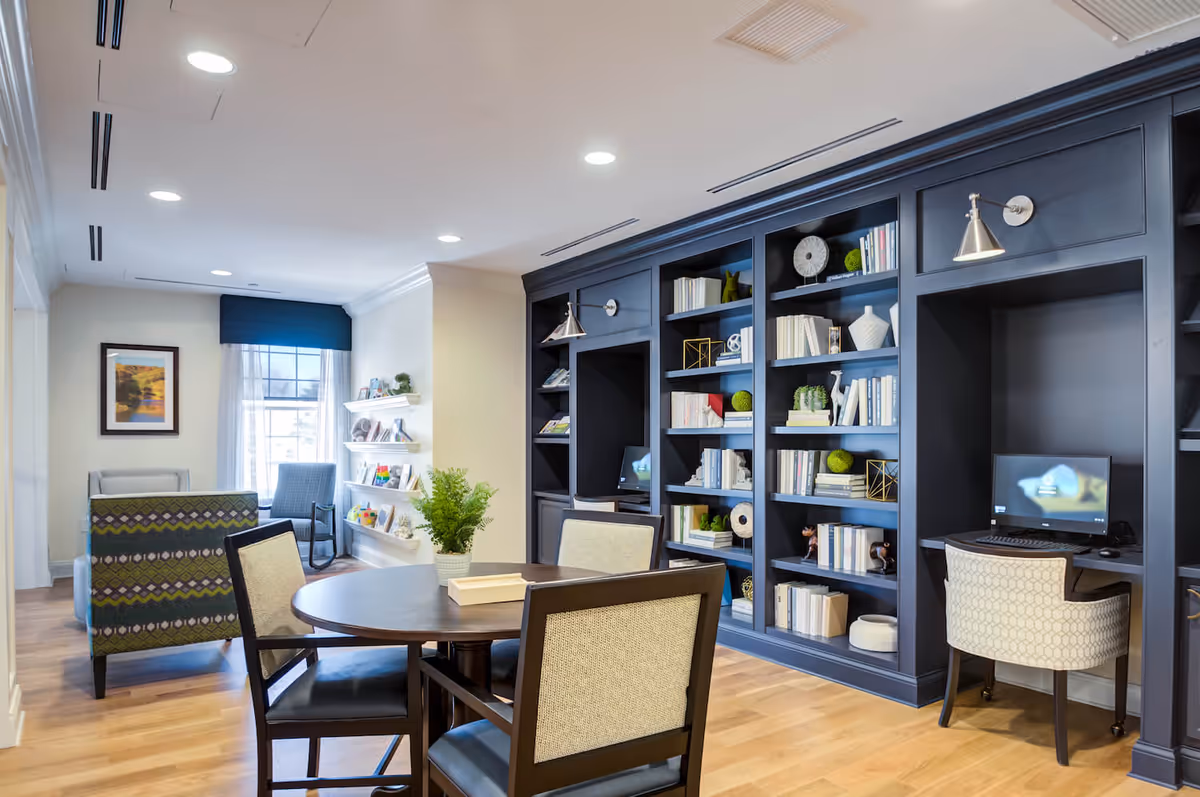 A well-lit common area with a round table and four chairs in the foreground. Behind the table is a large dark blue built-in bookshelf filled with books and decorative items. To the right, there is a small desk area with a computer and a cushioned chair. In the background, there are additional chairs near a window with sheer curtains and a framed picture on the wall. The floor is wooden, and the ceiling has recessed lighting.
